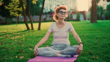 Parkta yoga. Nilüfer çiçekli orta yaşlı kadın yeşil çimlerin üzerinde yoga minderinde oturuyor. Sakinlik ve meditasyon kavramı. Yoga açık havada. Kıdemli kadın doğada meditasyon yapıyor ve yoga nilüfer pozisyonu uyguluyor.