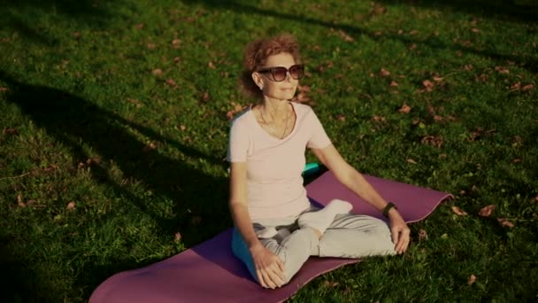 Mujer mayor meditando y ejercitando yoga posición de loto al aire libre. Mujer anciana haciendo ejercicios de estiramiento en estera de yoga en el parque en el césped verde al atardecer. Sensación de energía. Concepto calma y meditación — Vídeo de stock