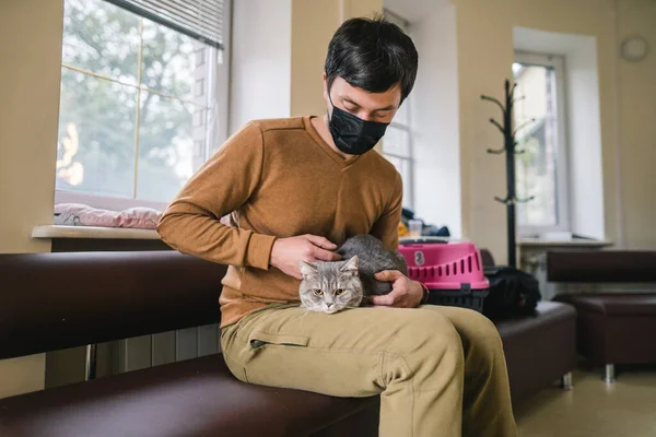 Man wearing protective mask with pet waiting in lobby for medical examination at veterinary clinic. Animal health. Owner of gray British cat waiting in lobby of animal clinic and is worried.