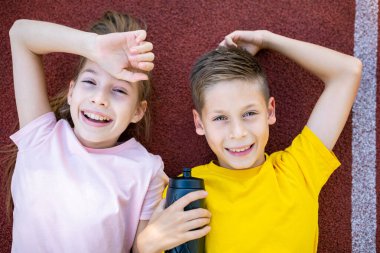 Mutlu spor çocukları erkek ve kız kardeş ikizler stadyumdaki koşu pistinde yatar, gülümser ve kameranın üst görüntüsüne bakarlar. Genç kız ve erkek atletler arka planda uzanmış stadyumda koşuyorlar ve gülümsüyorlar..