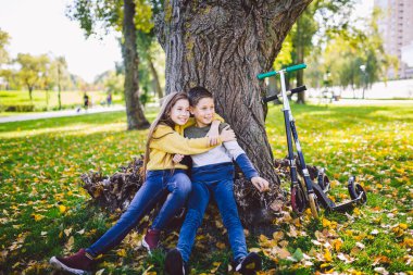 Çocuklar için aktif eğlence ve açık hava sporları. Mutlu bir çocukluk. Erkek ve kız ikizler bir sonbahar parkında motosiklet sürdükten sonra bir ağacın altında otururken mutlu bir şekilde poz veriyorlar. Merhaba sonbahar..