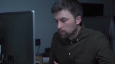Modern genç adam geceleri evden uzakta çalışıyor. Masaüstü bilgisayarında çalışan konsantre işadamı, klavyeye basıyor ve gece geç saatlerde ofiste monitör ekranına dikkatle bakıyor.