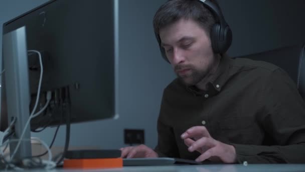 Muž na volné noze v košili, pracuje v noci doma u počítače. Nosí sluchátka, píše na klávesnici a dívá se na monitor. IT technik ve sluchátkách pracuje na počítači v kanceláři pozdě večer — Stock video
