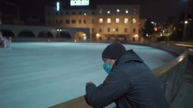 Kilitlenme teması yeni yıl tatili. Eğlence ve spor merkezleri kapandı. Noel tatilinde kapalı buz pateni pistinin yanında maske takan üzgün bir adam. Noel buz pateni pisti süslenmiş Noel ağacı boş