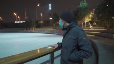 Maskeli erkek, akşam karantina, koronavirüs ve çovid salgını sırasında süslenmiş Noel ağacı ile buz pateni sahasının yanında duruyor. Geceleri buz pateni pistinde. Boş bir buz pateni sahası.