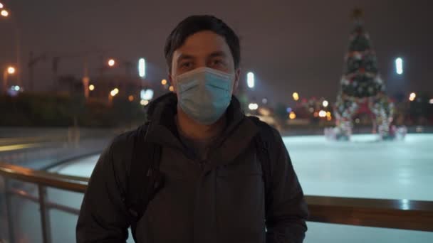 Smutny mężczyzna w masce patrzy na krytą lodową arenę z choinką w środku lodowiska wieczorem podczas zamknięcia, pandemii koronawirusowej. Puste miejsce publiczne na łyżwy — Wideo stockowe