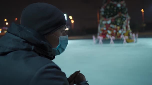 En ledsen man i mask tittar på en inomhusisarena med en julgran i mitten av rinken på kvällen under nedlåsning, coronavirus pandemi. Tomma offentliga plats för skridskoåkning — Stockvideo