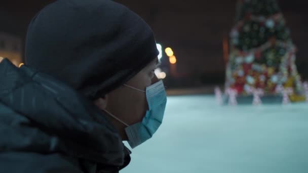 Hombre enmascarado se para cerca de pista de patinaje arena con árbol de Navidad decorado vacío durante la cuarentena, coronavirus y pandemia covid por la noche. Pista de patinaje por la noche. Una zona vacía de patinaje sobre hielo público — Vídeos de Stock