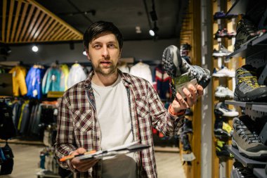 Satıcı, müşteriye açık hava mağazasında yürüyüş ayakkabısı seçimi konusunda tavsiye veriyor. Erkek turist dükkânı sahibi müşterilere yürüyüş botları denerken yardım ediyor. Genç satıcı kendinden emin bir şekilde dağ botu sunar. Sat, sat.