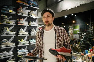 Satıcı, müşteriye açık hava mağazasında yürüyüş ayakkabısı seçimi konusunda tavsiye veriyor. Erkek turist dükkânı sahibi müşterilere yürüyüş botları denerken yardım ediyor. Genç satıcı kendinden emin bir şekilde dağ botu sunar. Sat, sat.