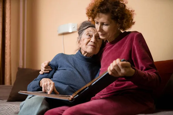 Mature daughter visits senior mother and has fun watching family photo album, sitting in embrace in living room couch. Mothers Day. Elderly woman and old gray hair grandmother laugh and remember youth