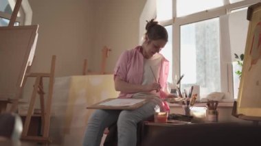 Beyaz kadın sanatçı, gün ışığında bir resim üzerinde çalışıyor. Mutlu ressam, atölyede fırça ve boyalarla bir sanat projesi çiziyor. Hobi olarak. Çalışan bir sanatçı. Yaratıcı meslek