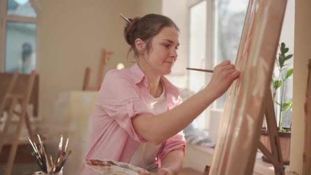 Fehér nő művész, aki egy festményen dolgozik fényes nappali stúdióban. Happy művész rajzol egy művészeti projekt festékek és ecsettel a műhelyben. Hobbi. Művész a munkahelyemen. Kreatív szakma — Stock videók