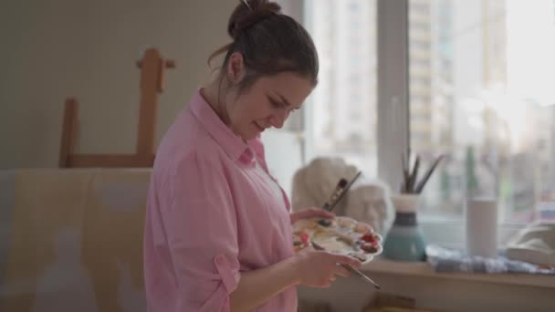 Leuke vrouw schildert op doek in een atelier. Kunstenaar die beeld schept. Kunstacademie of atelier. Werk met verf, penselen en ezel. Hobby en vrijetijdsconcept. Vrouwelijke schilder op het werk — Stockvideo