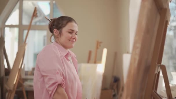 Leuke vrouw schildert op doek in een atelier. Kunstenaar die beeld schept. Kunstacademie of atelier. Werk met verf, penselen en ezel. Hobby en vrijetijdsconcept. Vrouwelijke schilder op het werk — Stockvideo