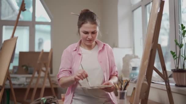 Beyaz kadın sanatçı, gün ışığında bir resim üzerinde çalışıyor. Mutlu ressam, atölyede fırça ve boyalarla bir sanat projesi çiziyor. Hobi olarak. Çalışan bir sanatçı. Yaratıcı meslek — Stok video