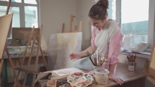 Söt kvinna målar på duk i en konstverkstad. Konstnär skapar bild. Konstskola eller ateljé. Arbeta med färger, penslar och staffli. Fritidskoncept och hobby. Kvinnlig målare på arbetsplatsen — Stockvideo