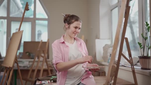 Femeia drăguță pictează pe pânză într-un atelier de artă. Artistul creează o imagine. Școala de artă sau studioul. Lucrați cu vopsele, perii și șevalet. Conceptul de hobby și petrecere a timpului liber. Femeie pictor la locul de muncă — Videoclip de stoc