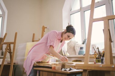 Sanat atölyesinde tuvale resim yapan hoş bir kadın. Ressam resim yaratıyor. Sanat okulu ya da stüdyo. Boya, fırça ve sehpayla çalış. Hobi ve eğlence konsepti. Çalışma alanındaki kadın ressam.
