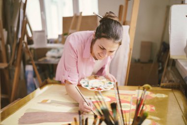 Sanat atölyesinde tuvale resim yapan hoş bir kadın. Ressam resim yaratıyor. Sanat okulu ya da stüdyo. Boya, fırça ve sehpayla çalış. Hobi ve eğlence konsepti. Çalışma alanındaki kadın ressam.