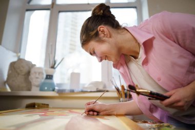 Beyaz kadın sanatçı, gün ışığında bir resim üzerinde çalışıyor. Mutlu ressam, atölyede fırça ve boyalarla bir sanat projesi çiziyor. Hobi olarak. Çalışan bir sanatçı. Yaratıcı meslek.