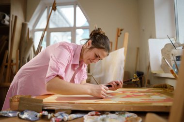 Sanat atölyesinde tuvale resim yapan hoş bir kadın. Ressam resim yaratıyor. Sanat okulu ya da stüdyo. Boya, fırça ve sehpayla çalış. Hobi ve eğlence konsepti. Çalışma alanındaki kadın ressam.