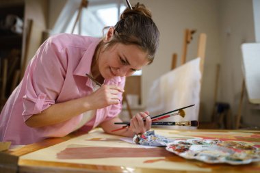 Sanat atölyesinde tuvale resim yapan hoş bir kadın. Ressam resim yaratıyor. Sanat okulu ya da stüdyo. Boya, fırça ve sehpayla çalış. Hobi ve eğlence konsepti. Çalışma alanındaki kadın ressam.