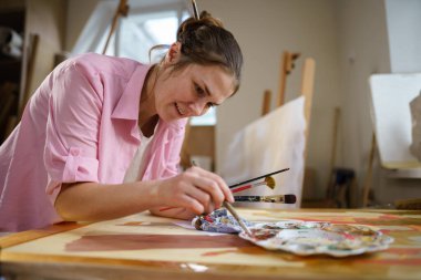 Sanat atölyesinde tuvale resim yapan hoş bir kadın. Ressam resim yaratıyor. Sanat okulu ya da stüdyo. Boya, fırça ve sehpayla çalış. Hobi ve eğlence konsepti. Çalışma alanındaki kadın ressam.
