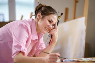Sanat atölyesinde tuvale resim yapan hoş bir kadın. Ressam resim yaratıyor. Sanat okulu ya da stüdyo. Boya, fırça ve sehpayla çalış. Hobi ve eğlence konsepti. Çalışma alanındaki kadın ressam.