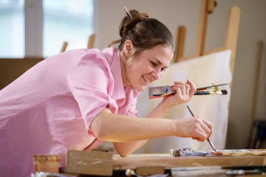 Sanat atölyesinde tuvale resim yapan hoş bir kadın. Ressam resim yaratıyor. Sanat okulu ya da stüdyo. Boya, fırça ve sehpayla çalış. Hobi ve eğlence konsepti. Çalışma alanındaki kadın ressam.