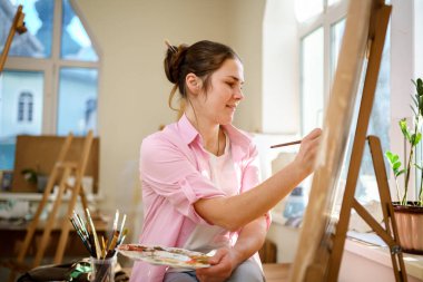 Beyaz kadın sanatçı, gün ışığında bir resim üzerinde çalışıyor. Mutlu ressam, atölyede fırça ve boyalarla bir sanat projesi çiziyor. Hobi olarak. Çalışan bir sanatçı. Yaratıcı meslek.