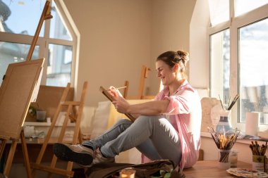 Beyaz kadın sanatçı, gün ışığında bir resim üzerinde çalışıyor. Mutlu ressam, atölyede fırça ve boyalarla bir sanat projesi çiziyor. Hobi olarak. Çalışan bir sanatçı. Yaratıcı meslek.