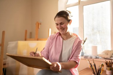 Sanat atölyesinde tuvale resim yapan hoş bir kadın. Ressam resim yaratıyor. Sanat okulu ya da stüdyo. Boya, fırça ve sehpayla çalış. Hobi ve eğlence konsepti. Çalışma alanındaki kadın ressam.