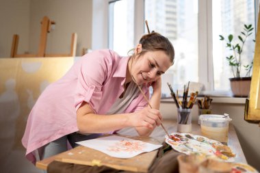 Sanat atölyesinde tuvale resim yapan hoş bir kadın. Ressam resim yaratıyor. Sanat okulu ya da stüdyo. Boya, fırça ve sehpayla çalış. Hobi ve eğlence konsepti. Çalışma alanındaki kadın ressam.