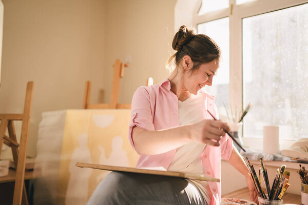 Кавказская художница работает над картиной в ярко-дневной студии. Счастливый художник рисует художественный проект с красками и кистью в мастерской. Хобби. Художник за работой. Творческая профессия.