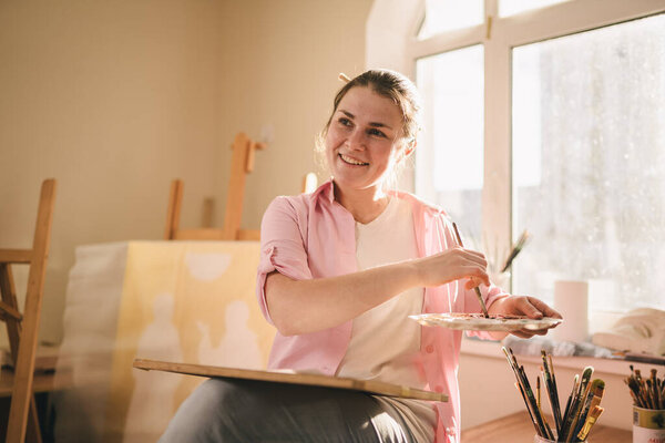 Милая женщина рисует на холсте в художественной мастерской. Художник создает картину. Школа искусств или студия. Работа с красками, кисточками и мольбертом. Концепция хобби и отдыха. Художница на рабочем месте.