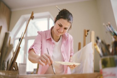 Beyaz kadın sanatçı, gün ışığında bir resim üzerinde çalışıyor. Mutlu ressam, atölyede fırça ve boyalarla bir sanat projesi çiziyor. Hobi olarak. Çalışan bir sanatçı. Yaratıcı meslek.