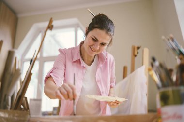 Sanat atölyesinde tuvale resim yapan hoş bir kadın. Ressam resim yaratıyor. Sanat okulu ya da stüdyo. Boya, fırça ve sehpayla çalış. Hobi ve eğlence konsepti. Çalışma alanındaki kadın ressam.