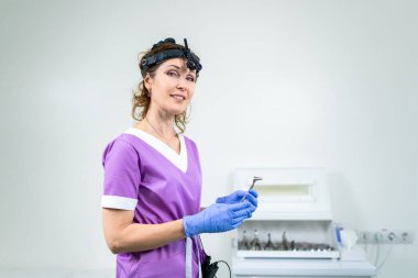 KBB kliniğinde çalışan orta yaşlı, mor üniformalı beyaz kadın muayene odasındaki kameraya bakıyor. Hastanede uzman bir otolaryngolog..