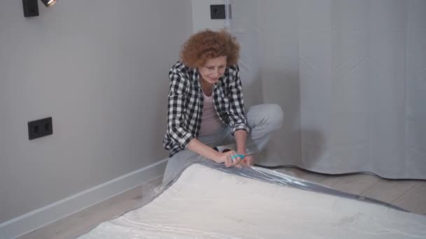 Happy adult woman unpacks new orthopedic mattress. Unpacking mattress with scissors pressed into bag. Moving to new apartment. New house without furniture. Elderly female opens mattress at home — Stock Video