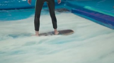 Genç, sörf tahtasında dalga simülatöründe kapalı alanda sörf yapıyor. Genç sörfçü, üretilmiş dalgalar üzerine eğitim alırken. Su sporu aktivitesi. Sörfçü çocuk sörf taklitçisi olarak sörf yapmayı seviyor. Sörf eğitimi