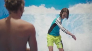 Genç, sörf tahtasında dalga simülatöründe kapalı alanda sörf yapıyor. Genç sörfçü, üretilmiş dalgalar üzerine eğitim alırken. Su sporu aktivitesi. Sörfçü çocuk sörf taklitçisi olarak sörf yapmayı seviyor. Sörf eğitimi