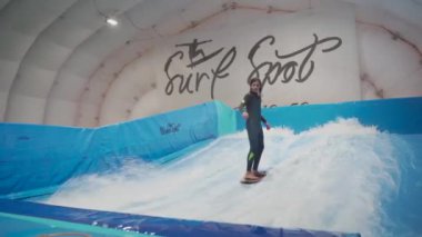 Genç, sörf tahtasında dalga simülatöründe kapalı alanda sörf yapıyor. Genç sörfçü, üretilmiş dalgalar üzerine eğitim alırken. Su sporu aktivitesi. Sörfçü çocuk sörf taklitçisi olarak sörf yapmayı seviyor. Sörf eğitimi