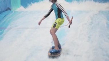 Genç, sörf tahtasında dalga simülatöründe kapalı alanda sörf yapıyor. Genç sörfçü, üretilmiş dalgalar üzerine eğitim alırken. Su sporu aktivitesi. Sörfçü çocuk sörf taklitçisi olarak sörf yapmayı seviyor. Sörf eğitimi
