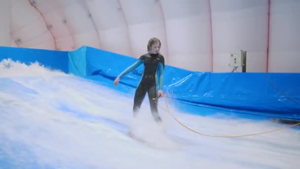 Adolescente está montando prancha de surf no simulador de ondas dentro de casa. Surfista jovem durante o treino em ondas geradas. Actividades desportivas aquáticas. Surfista garoto gosta de surfar no imitador de surf indoor. Treinamento de surf — Vídeo de Stock