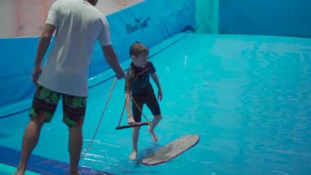 Actividad deportiva acuática, concepto de equilibrio. Pequeño atleta disfrutando del surf indoor. Los paseos adolescentes abordan olas en el simulador en el complejo deportivo. Entrenador de surf y estudiante en sesión en simulador de ola — Vídeo de stock