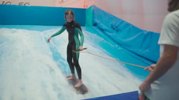 Actividad deportiva acuática, concepto de equilibrio. Pequeño atleta disfrutando del surf indoor. Los paseos adolescentes abordan olas en el simulador en el complejo deportivo. Entrenador de surf y estudiante en sesión en simulador de ola — Vídeos de Stock