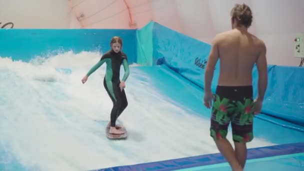Water sport activity, balance concept. Little athlete enjoying indoor surfing. Teenager rides board on waves on simulator in sports complex. Surfing coach and student in session on wave simulator — Stock Video