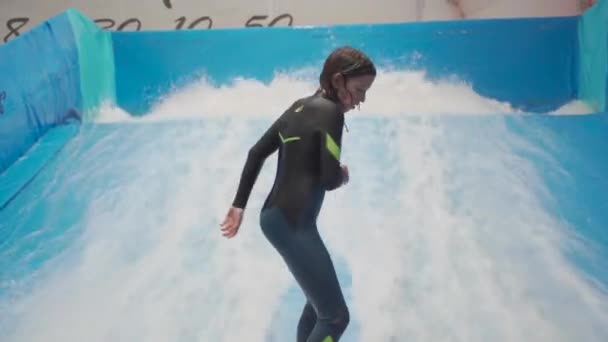 Genç, sörf tahtasında dalga simülatöründe kapalı alanda sörf yapıyor. Genç sörfçü, üretilmiş dalgalar üzerine eğitim alırken. Su sporu aktivitesi. Sörfçü çocuk sörf taklitçisi olarak sörf yapmayı seviyor. Sörf eğitimi — Stok video