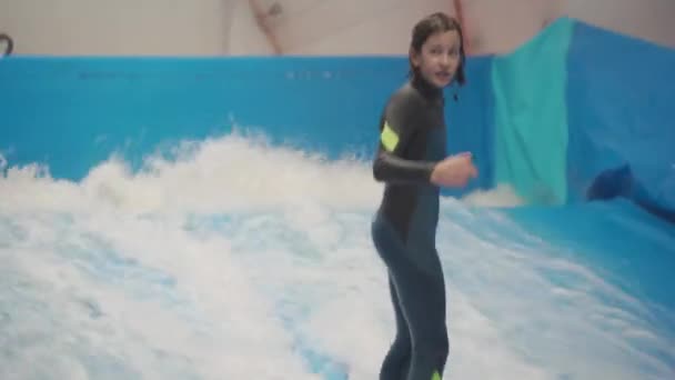 Sporty wodne, koncepcja równowagi. Mały sportowiec cieszący się krytym surfingiem. Nastolatek jeździ deską na falach na symulatorze w kompleksie sportowym. Trener surfingu i student w sesji na symulatorze fali — Wideo stockowe